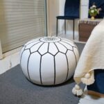 White Moroccan leather pouf with black embroidered stitching placed in a cozy bedroom setting